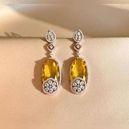 Drop earrings with elongated yellow glass stones and pavé American diamond floral accents, set in a halo frame, displayed on a satin nude background.